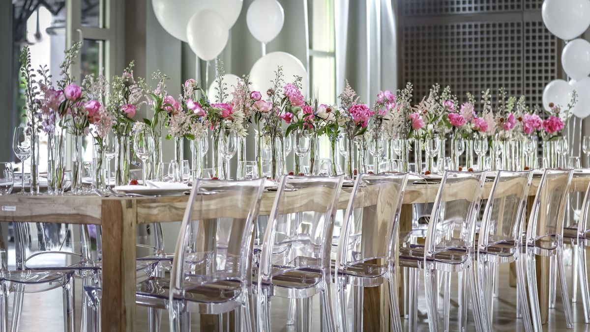 Ansicht von der Seite auf einen langen hölzernen Tisch mit vielen Blumenvasen, Luftballons und durchsichtigen Stühlen in der Eventlocation München