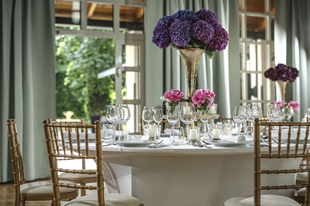 Ansicht von der Seite auf einen runden Tisch in einem Speisesaal mit rosa und violetten Blumen und goldenen Stühlen in der Eventlocation München